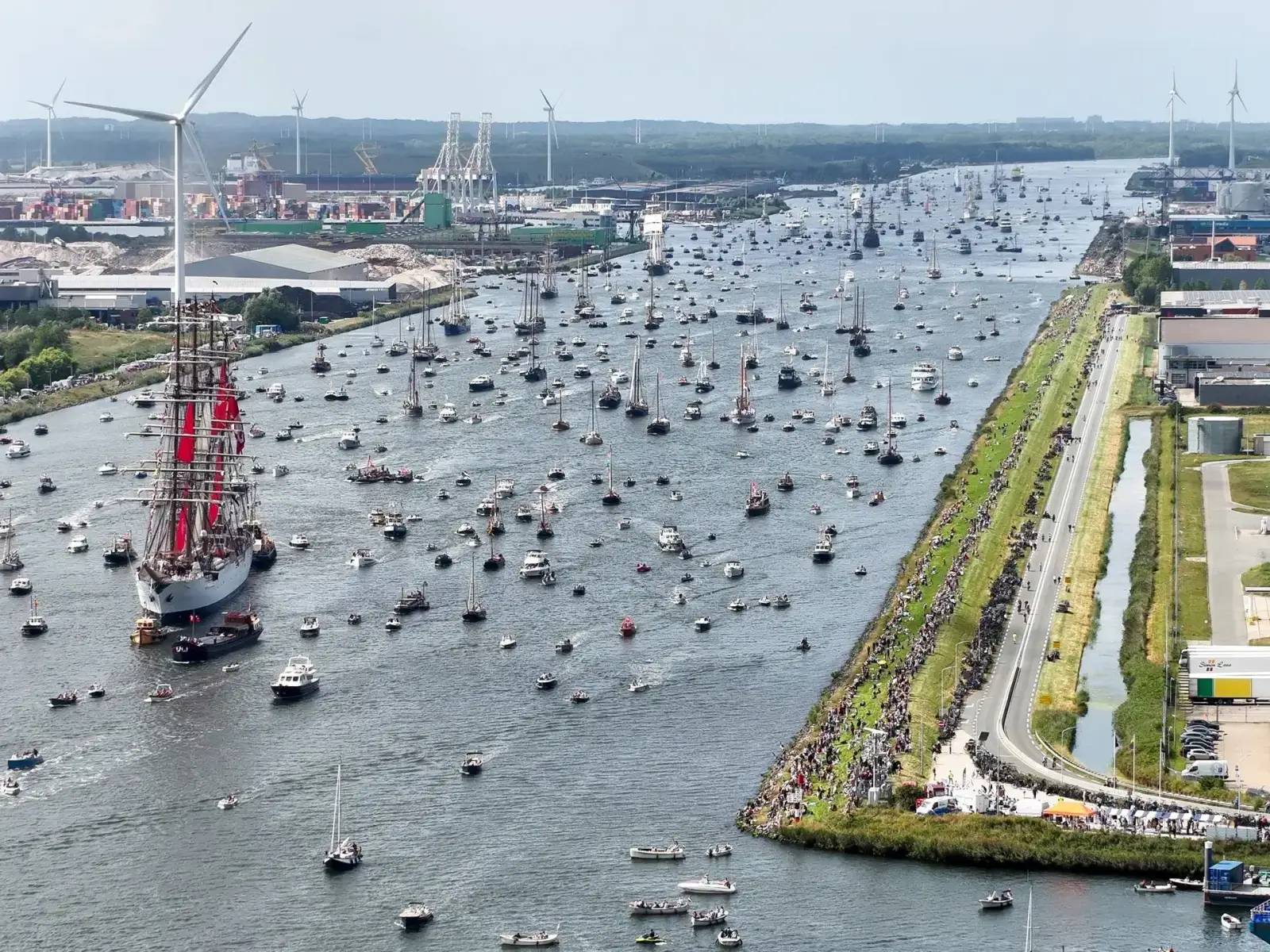 Tallship met rode zeilen tussen honderden boten, met publiek op de dijk