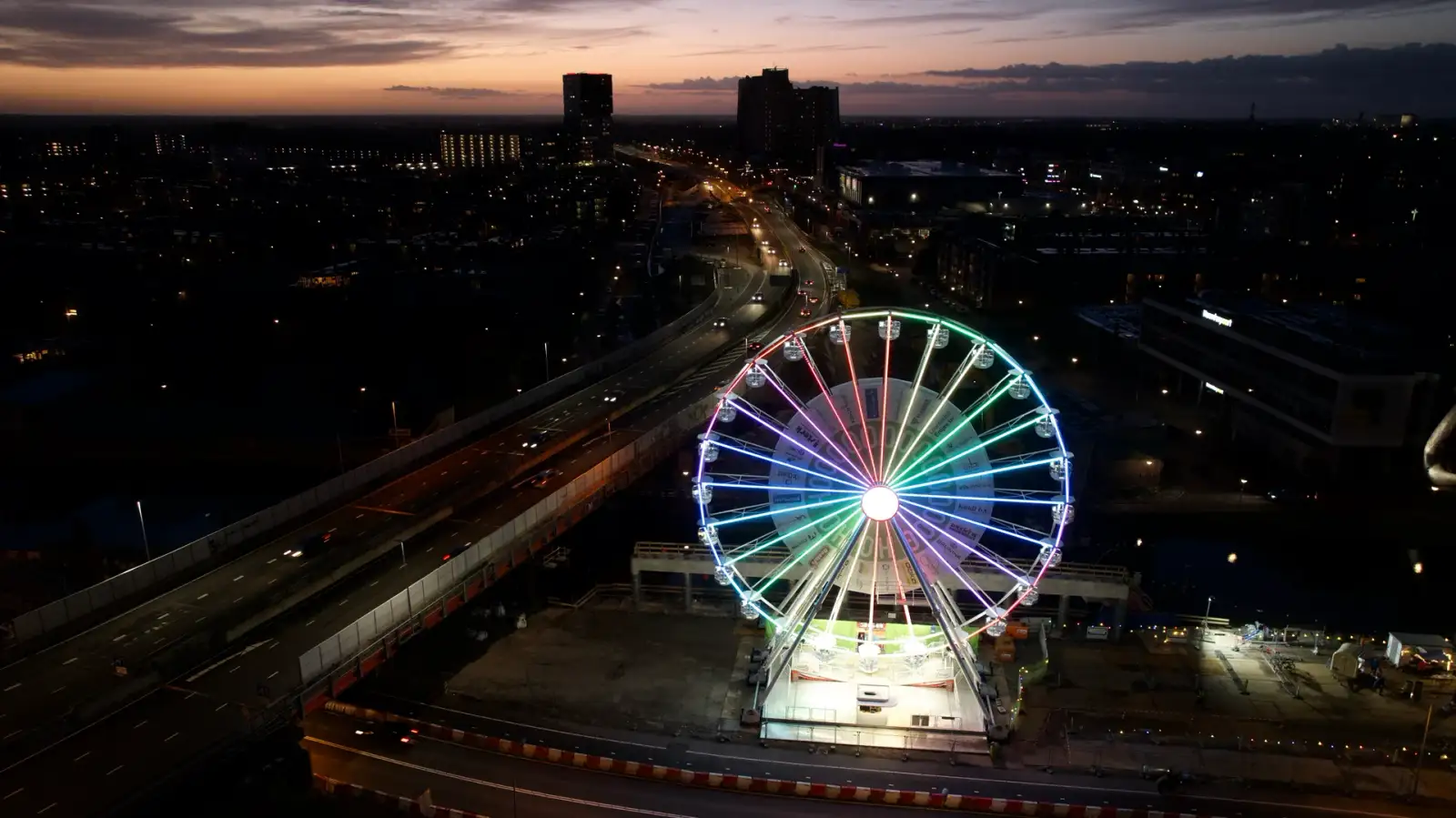 Verlicht reuzenrad bij nacht naast een drukke snelweg, vanuit de lucht