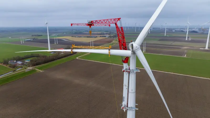 Windturbine met een rode kraan ernaast tijdens onderhoud, tussen akkers